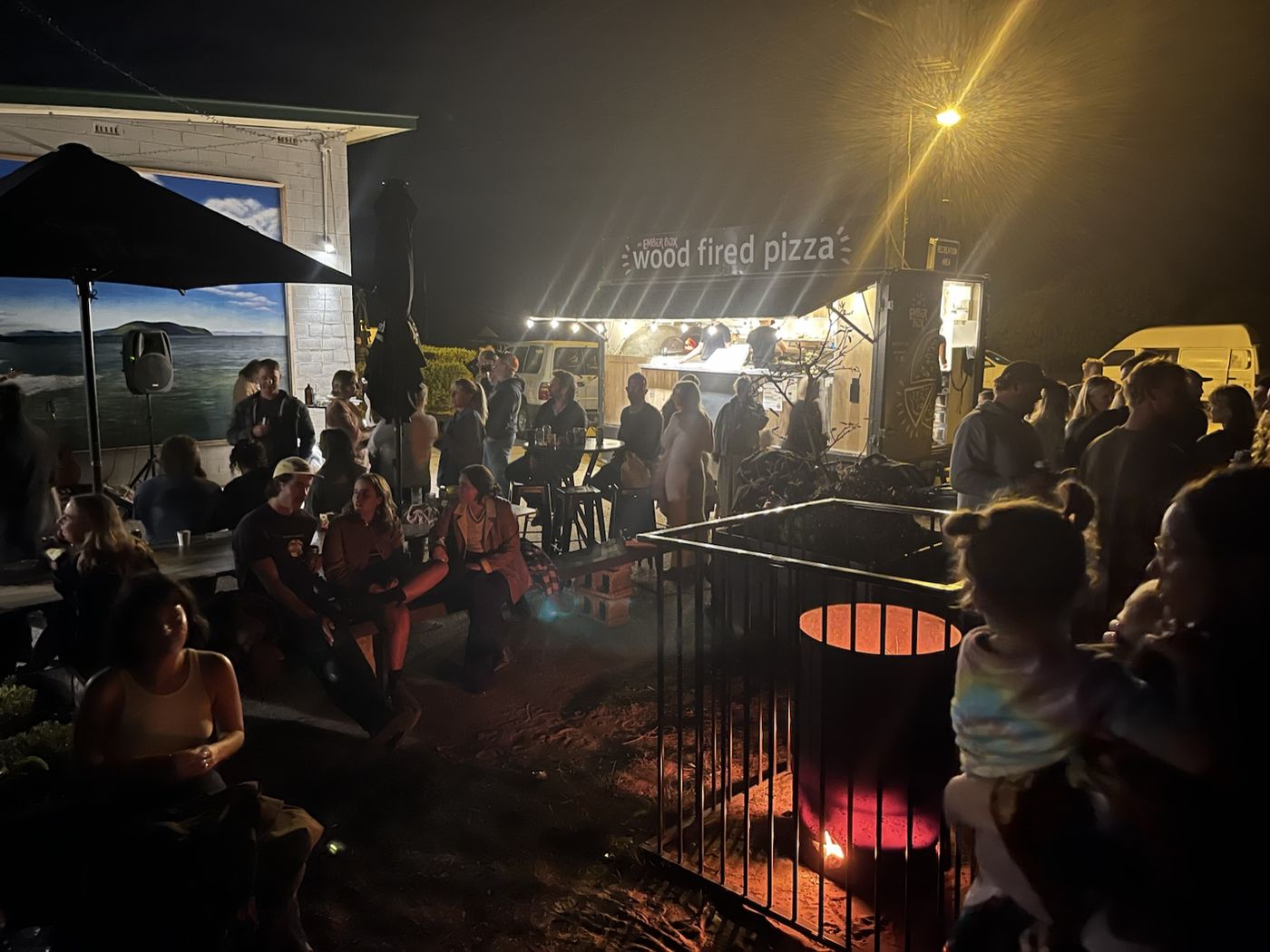 Guests gathered around a fire pit at night in the beer garden by the Ember Bus
